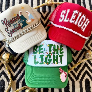 May include: Three Christmas themed trucker hats. The first hat is beige with a gold chain trim and the text "Reason for the Season". The second hat is red with white pearl trim and the text "Sleigh". The third hat is green with white string lights and the text "Be the Light".