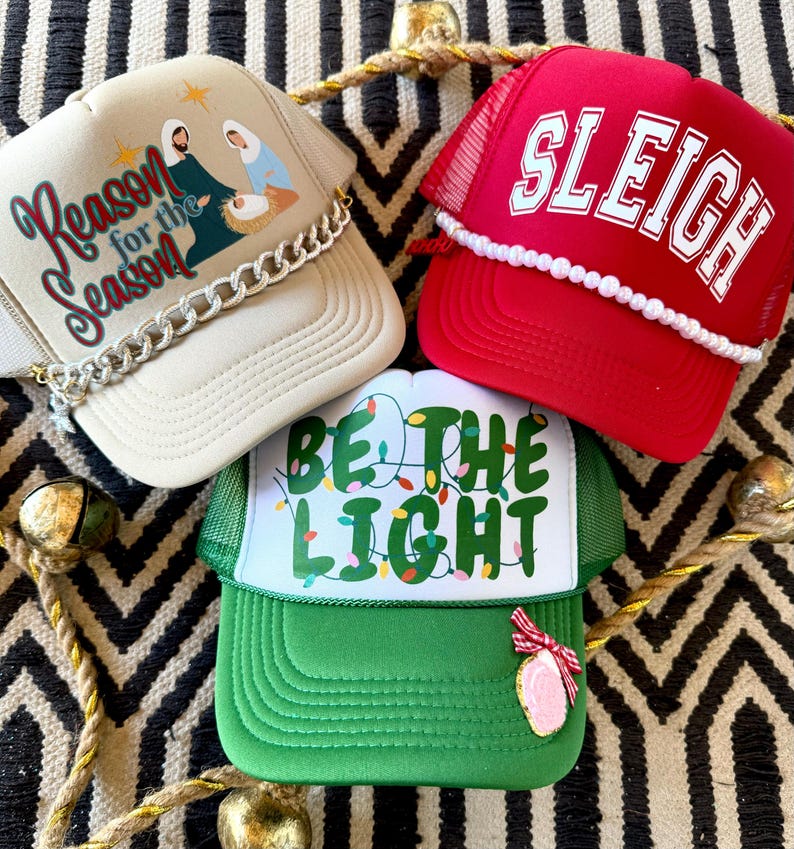 May include: Three Christmas themed trucker hats. The first hat is beige with a gold chain trim and the text "Reason for the Season". The second hat is red with white pearl trim and the text "Sleigh". The third hat is green with white string lights and the text "Be the Light".