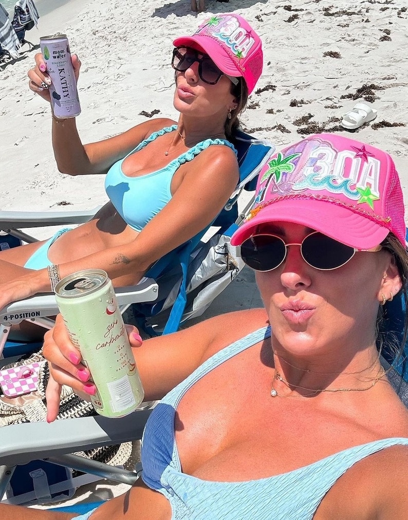 Op de afbeelding: Twee vrouwen in blauwe en witte bikini's en roze baseballpetten met "30A" erop geborduurd. Een vrouw houdt een blikje bruisend water vast en de andere een blikje koolwaterstofvrij kokoswater.