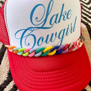 以下が含まれることがあります： 赤と白のトラッカーハットに、青い刺繍で「Lake Cowgirl」と書いてあります。ハットのつばにはカラフルなプラスチックチェーンが付いています。