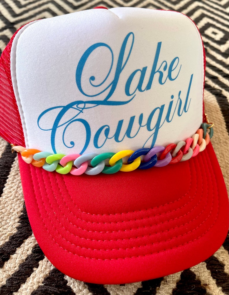 以下が含まれることがあります： 赤と白のトラッカーハットに、青い刺繍で「Lake Cowgirl」と書いてあります。ハットのつばにはカラフルなプラスチックチェーンが付いています。