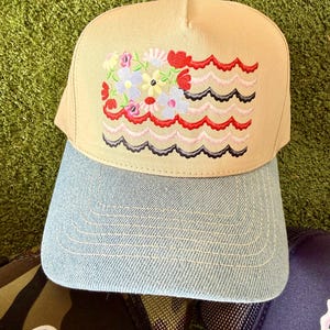 May include: Tan baseball cap with a light blue denim brim. The cap features an embroidered design of colorful flowers and wavy lines. The cap has a light blue button on top.