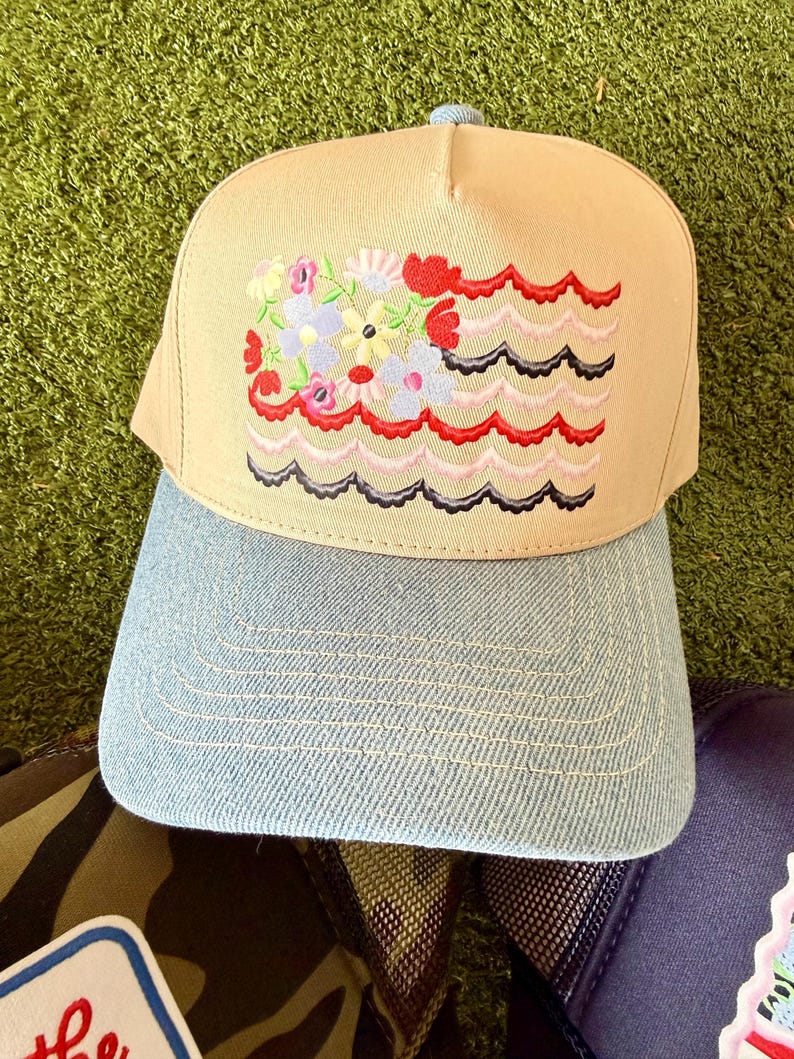 May include: Tan baseball cap with a light blue denim brim. The cap features an embroidered design of colorful flowers and wavy lines. The cap has a light blue button on top.