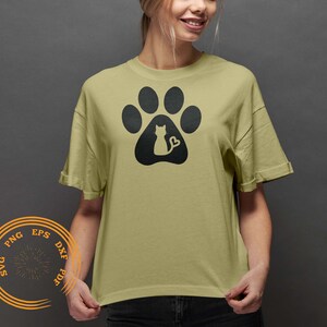 K&ouml;nnte beinhalten: Ein hellgr&uuml;nes T-Shirt mit einem schwarzen Pfotenabdruck-Design. Der Pfotenabdruck hat eine Silhouette einer Katze, die darin sitzt, mit einem herzf&ouml;rmigen Schwanz.