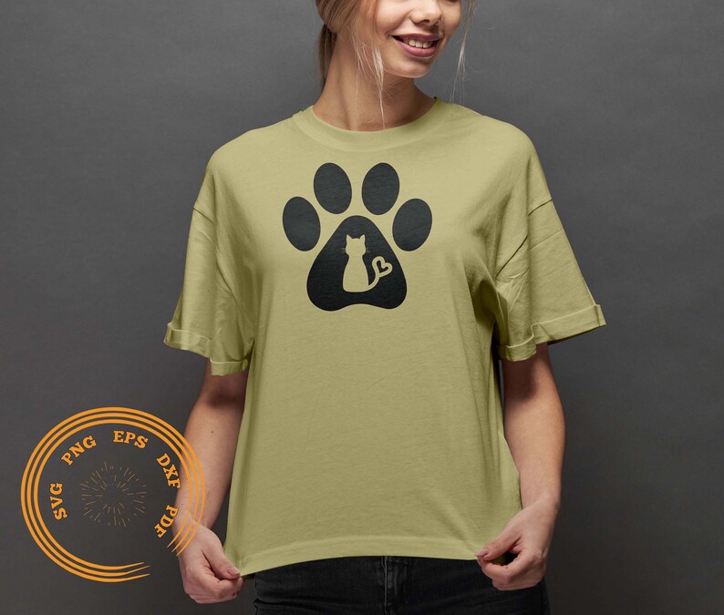 K&ouml;nnte beinhalten: Ein hellgr&uuml;nes T-Shirt mit einem schwarzen Pfotenabdruck-Design. Der Pfotenabdruck hat eine Silhouette einer Katze, die darin sitzt, mit einem herzf&ouml;rmigen Schwanz.