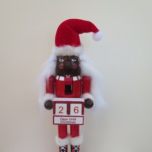 May include: A wooden Christmas nutcracker with a red and white Santa suit, black boots, and glasses. The nutcracker is holding a sign that says "26 Days Until Christmas".