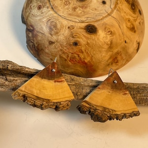 May include: A pair of wooden earrings in the shape of a triangle. The earrings are made from a light brown wood with dark brown accents. The earrings are attached to silver hooks.