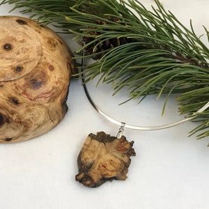 Puede incluir: Un collar choker plateado con un colgante de madera. El colgante es una pieza de madera natural con un patrón de veta único. El collar se muestra sobre una superficie blanca con un cuenco de madera y ramas de pino.