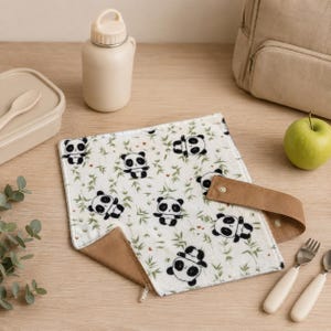 Könnte beinhalten: Ein quadratisches Stofftuch mit weißem Hintergrund und einem wiederkehrenden Muster aus schwarz-weißen Pandas und grünen Bambuszweigen. Das Tuch hat einen braunen Stoffrand, einen Metallreißverschluss und einen braunen Riemen mit Druckknopf.