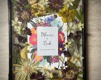 May include: A framed pressed flower arrangement with a white card inside. The card has the text "Mhairi + Nick" and "3rd September 2022".