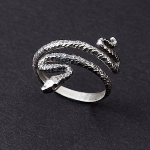 Op de afbeelding: Een zilverkleurige ring in de vorm van een slang, gewikkeld rond de vinger. Het lichaam van de slang heeft een textuur met schubbenpatroon en de kop is gedetailleerd met een kleine, open mond. De ring staat tegen een donkere achtergrond.