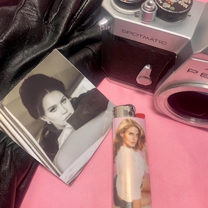 May include: A collection of items including a vintage silver Spotmatic camera, a black leather glove, a photograph, and a lighter with a woman's image. The lighter is white and pink. The photograph is black and white.