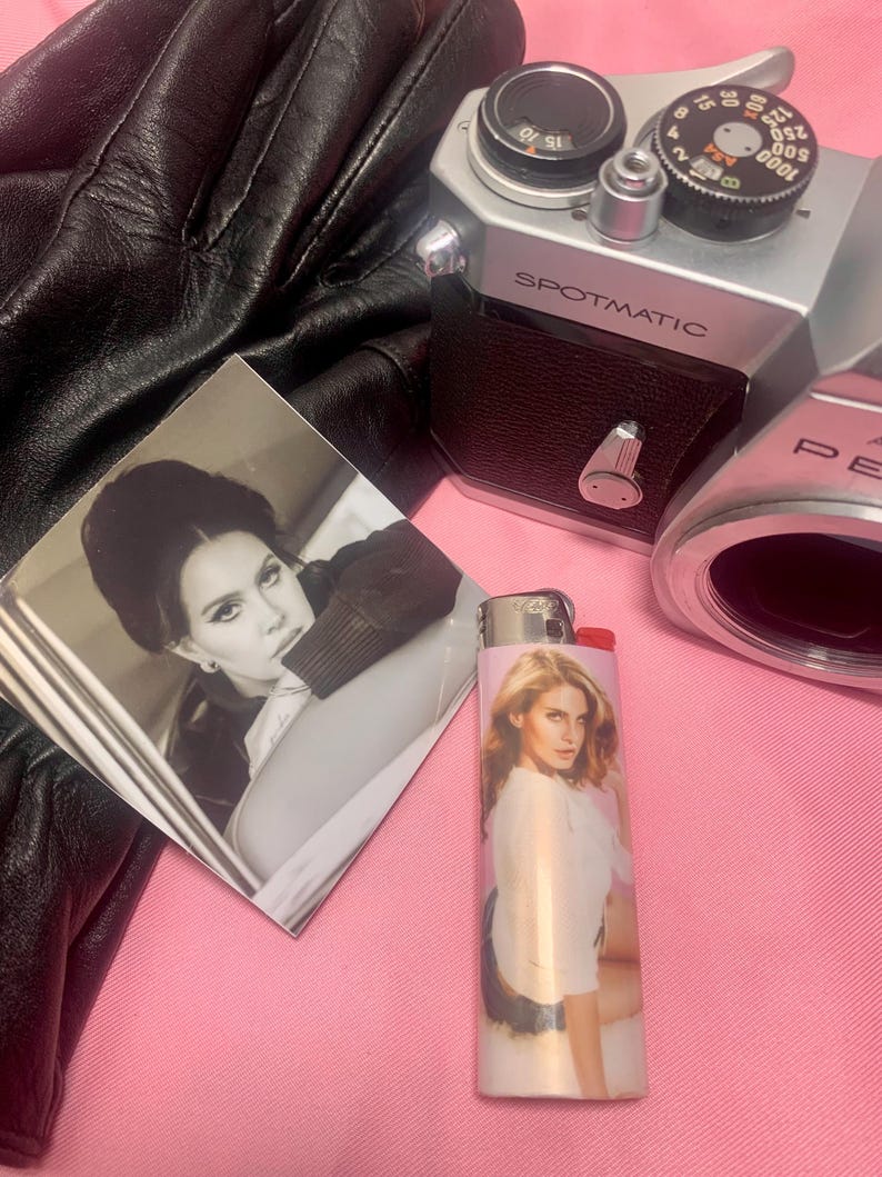 May include: A collection of items including a vintage silver Spotmatic camera, a black leather glove, a photograph, and a lighter with a woman's image. The lighter is white and pink. The photograph is black and white.