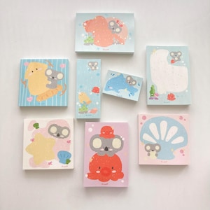 May include: Set of 8 colorful sticky notes featuring cute koala illustrations with various ocean-themed designs. The notes include a pink coral reef, a blue ocean wave, a yellow seashell, a blue dolphin, a white seashell, a blue and white striped background, a yellow and blue striped background, and a red octopus.