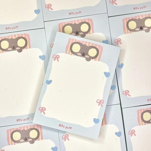 以下が含まれることがあります： 青いと白いメモ帳の束。コアラのイラストが描かれており、コアラはピンクと黄色のストライプのベッドで寝ています。メモ帳はピンクとブルーの縁取りで、ハートとリボンが付いています。