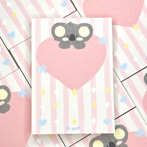 以下が含まれることがあります： ピンクのハートの上にグレーのコアラが描かれた長方形のメモ帳。背景にはピンクと白のストライプがあり、青いハート、黄色の星、そして「Po puff」という文字が散りばめられています。