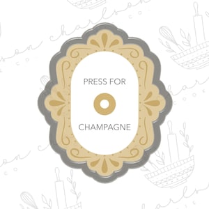 Może przedstawiać: Dekoracyjna foremka do ciasteczek w kolorze szarym i złotym z ozdobnym ramieniem. Foremka ma kształt ramki do obrazu i zawiera napis "Press for Champagne" wewnątrz.