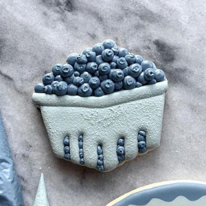 Puede incluir: Una galleta de azúcar azul y blanca decorada con una cesta de arándanos. La galleta es de color azul claro y los arándanos son de un azul más oscuro. La cesta está hecha de glaseado blanco y tiene una superficie texturizada.