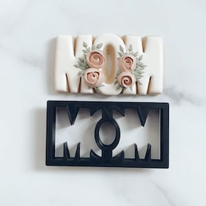 May include: A white cookie decorated with pink and green frosting in the shape of the word "MOM". A black plastic cookie cutter in the shape of the word "MOM" is also shown.