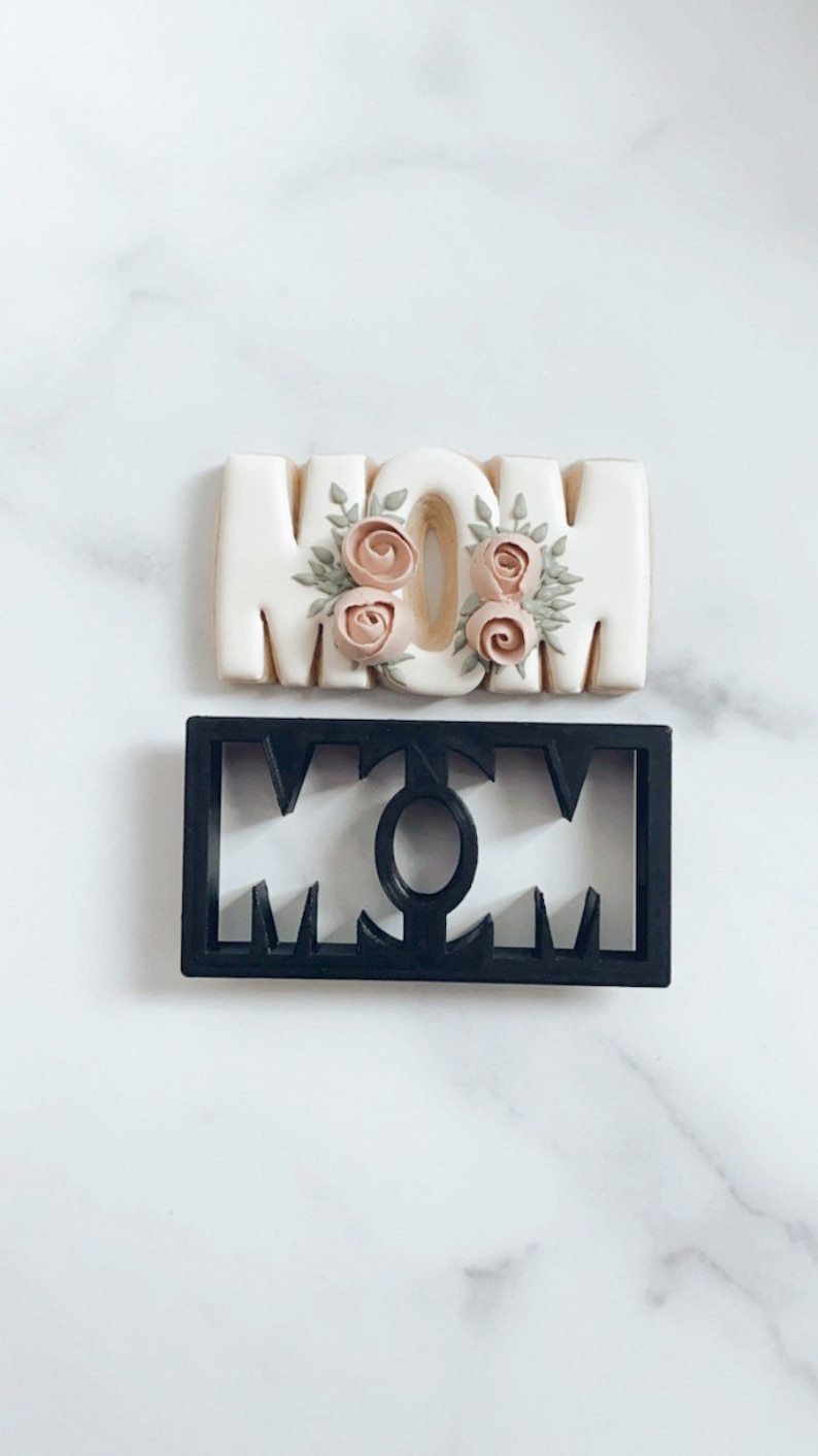 May include: A white cookie decorated with pink and green frosting in the shape of the word "MOM". A black plastic cookie cutter in the shape of the word "MOM" is also shown.