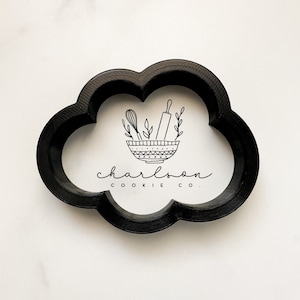 Może przedstawiać: Czarny wykrawacz do ciastek w kształcie chmury z białym wnętrzem. Wnętrze przedstawia miskę z narzędziami do pieczenia i liśćmi, a pod spodem napis "charlson cookie co.". Wykrawacz znajduje się na białym tle.