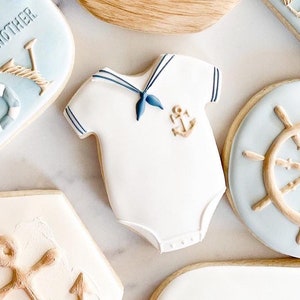 May include: A set of nautical themed sugar cookies decorated with blue and white icing. The cookies feature a sailboat, an anchor, a steering wheel, and a baby onesie with a sailor collar. The cookies are decorated with gold accents.