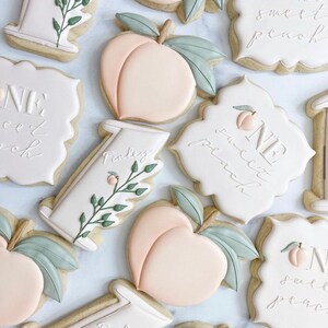 May include: A set of decorated sugar cookies with peach and leaf designs. The cookies are decorated with white icing and the text "One Sweet Peach" and "Finley".