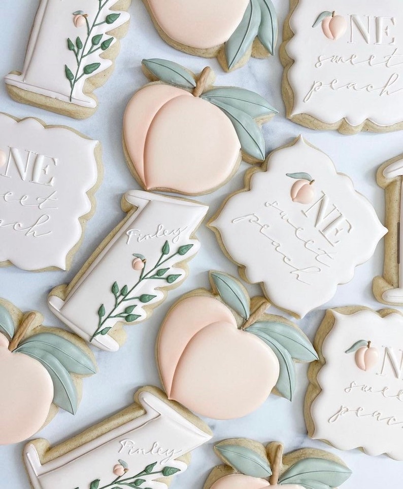 May include: A set of decorated sugar cookies with peach and leaf designs. The cookies are decorated with white icing and the text "One Sweet Peach" and "Finley".
