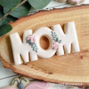 May include: A set of three pink sugar cookies decorated with white icing and floral details. The cookies spell out the word "MOM".