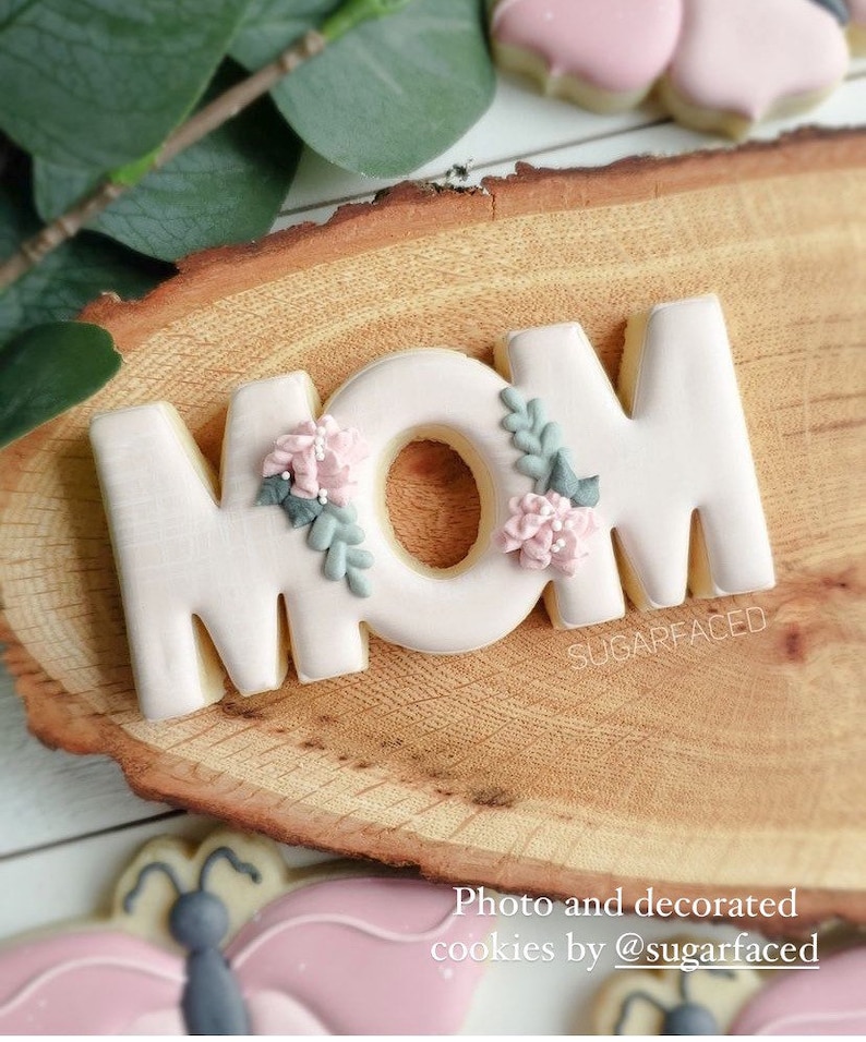 May include: A set of three pink sugar cookies decorated with white icing and floral details. The cookies spell out the word "MOM".
