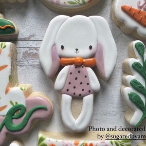 May include: A set of decorated sugar cookies featuring a bunny, a leaf, a number one, and a cookie with a carrot and green leaves. The bunny is white with pink ears and a pink and black polka dot dress. The leaf is green and the number one is white with orange carrots and green leaves. The cookie with the carrot and green leaves is white with an orange carrot and green leaves.
