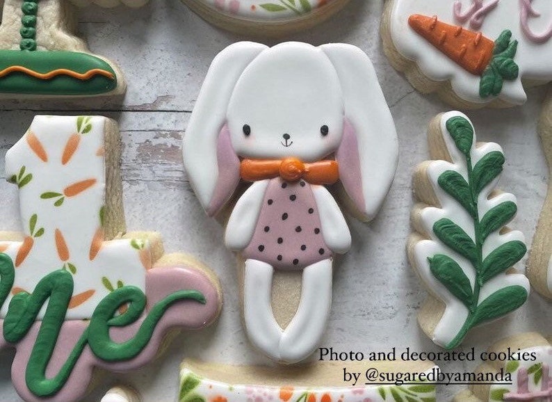 May include: A set of decorated sugar cookies featuring a bunny, a leaf, a number one, and a cookie with a carrot and green leaves. The bunny is white with pink ears and a pink and black polka dot dress. The leaf is green and the number one is white with orange carrots and green leaves. The cookie with the carrot and green leaves is white with an orange carrot and green leaves.