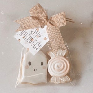 Puede incluir: Una bolsa de plástico transparente llena de dos galletas de azúcar con temática de Halloween. Una galleta es un fantasma con una cara sonriente y la otra es un bastón de caramelo. La bolsa está atada con una cinta de arpillera marrón y una etiqueta que dice "Feliz Halloween".