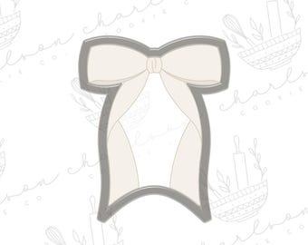 May include: A gray metal cookie cutter in the shape of a bow. The bow is tied with a white ribbon.