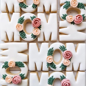 May include: Decorated sugar cookies in the shape of the word "MOM" with green leaves and pink and peach flowers. Photo and decorated cookies by @wellingtonbakehouse.