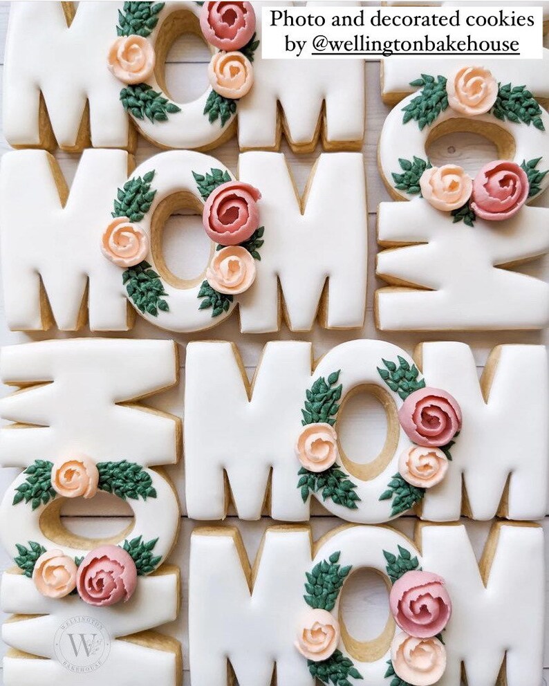 May include: Decorated sugar cookies in the shape of the word "MOM" with green leaves and pink and peach flowers. Photo and decorated cookies by @wellingtonbakehouse.