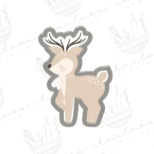 Puede incluir: Un cortador de galletas de metal gris en forma de ciervo con manchas marrones. El ciervo tiene ojos grandes y una pajarita.