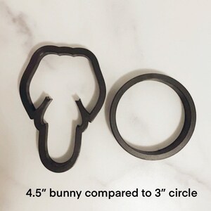 May include: A black 4.5 inch bunny cookie cutter next to a 3 inch black circle cookie cutter. The cookie cutters are on a white surface.
