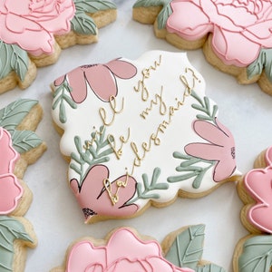 May include: A set of four decorated sugar cookies with floral designs. One cookie features the text "Will you be my bridesmaid?" in gold lettering.