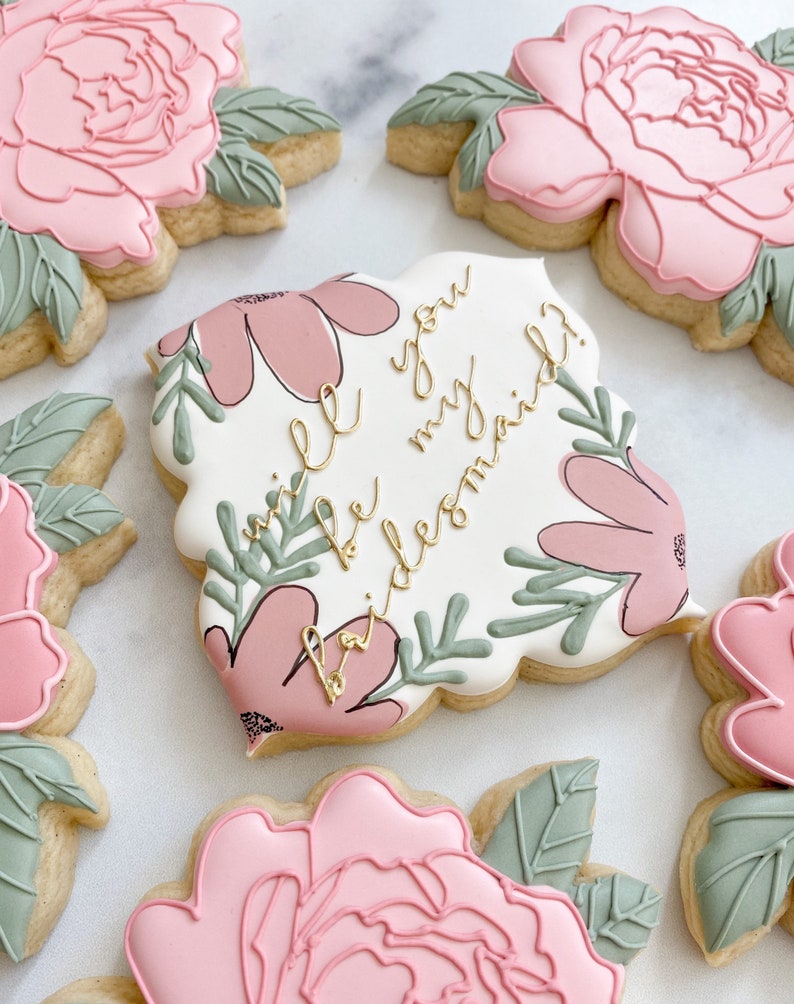 May include: A set of four decorated sugar cookies with floral designs. One cookie features the text "Will you be my bridesmaid?" in gold lettering.