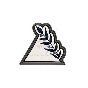May include: A gray triangle cookie cutter with a dark green leafy design. The cutter has a double outline and the text "cookie co." at the bottom.