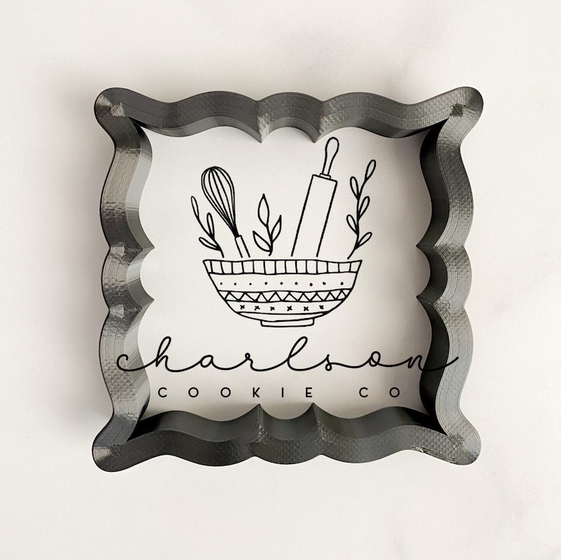 May include: A black cookie cutter in the shape of a square with rounded corners. The cutter features a design of a bowl with a rolling pin, whisk, and leaves inside. The text "charlson cookie co" is below the bowl.