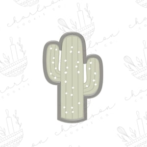Puede incluir: Un cortador de galletas de metal gris en forma de cactus con un diseño verde y blanco. El cactus tiene un cuerpo alto y delgado con múltiples brazos y puntos blancos.