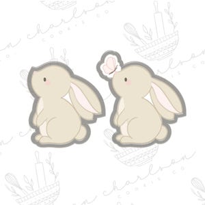 May include: Two bunny-shaped cookie cutters. One bunny is plain, the other has a butterfly above its nose. Both are light beige with pink accents and a gray outline. The background is white with a faint pattern of kitchen tools.