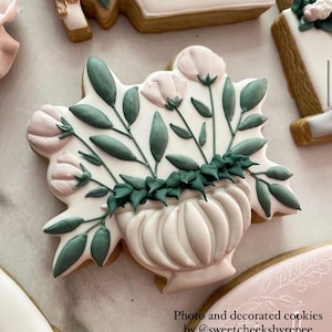 May include: A decorated sugar cookie with a white icing base and a design of a white pot with green leaves and pink flowers. The cookie is on a white surface. Photo and decorated cookies by @sweetcheeksbyrenee