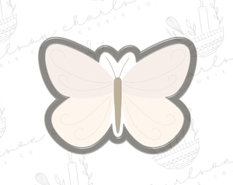 May include: A butterfly-shaped cookie cutter with a light gray outline. The butterfly's wings are a soft cream color with delicate swirl details. The body is a light brown color. The background is white with a repeating pattern of kitchen utensils.