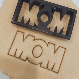 May include: A black plastic cookie cutter in the shape of the word "MOM" is on top of a piece of cookie dough with the word "MOM" cut out.