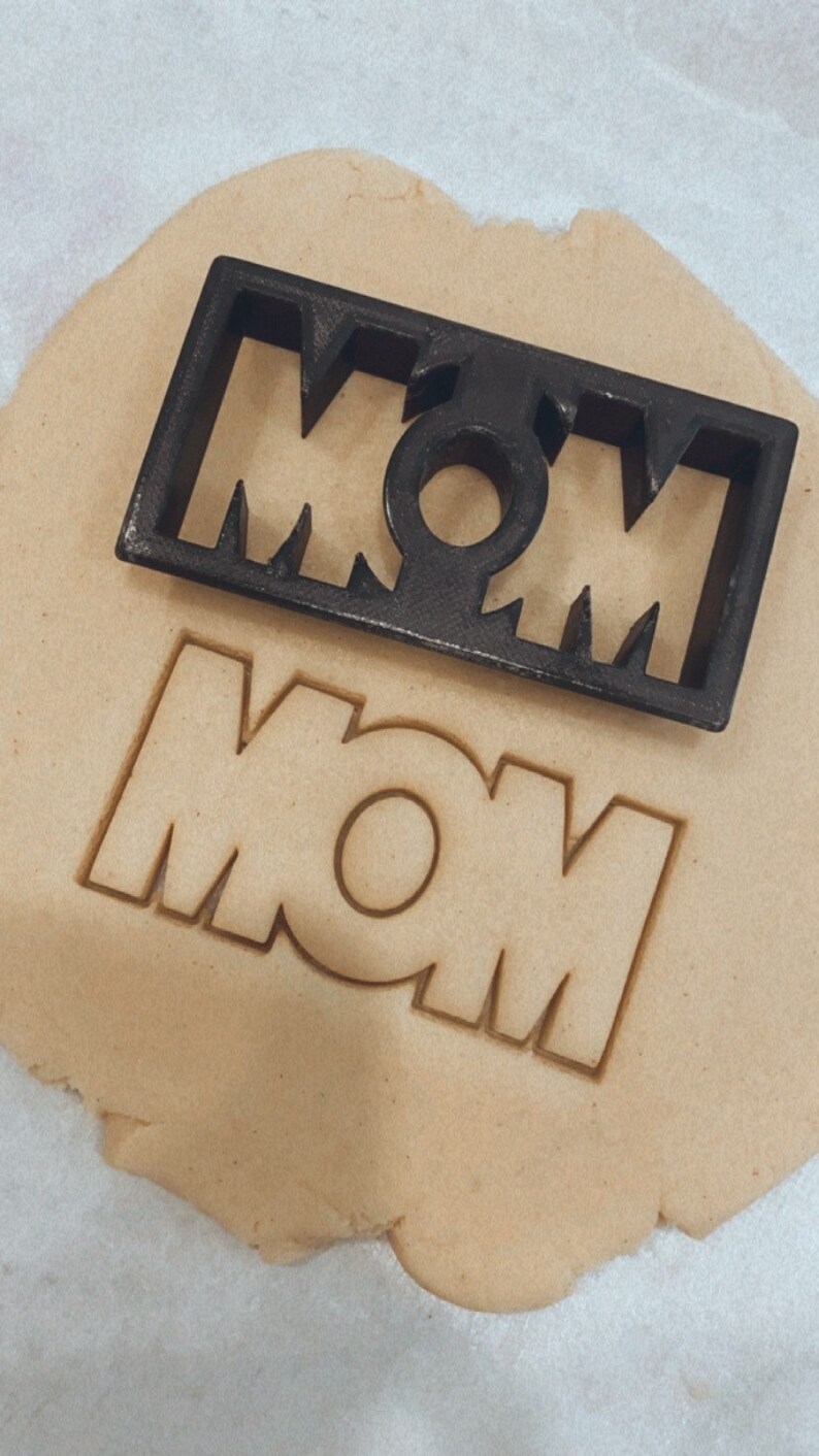 May include: A black plastic cookie cutter in the shape of the word "MOM" is on top of a piece of cookie dough with the word "MOM" cut out.