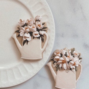 May include: Two white sugar cookies decorated with white icing and a floral design. The cookies are shaped like watering cans and are filled with white flowers. The cookies are on a white plate and a white marble surface.