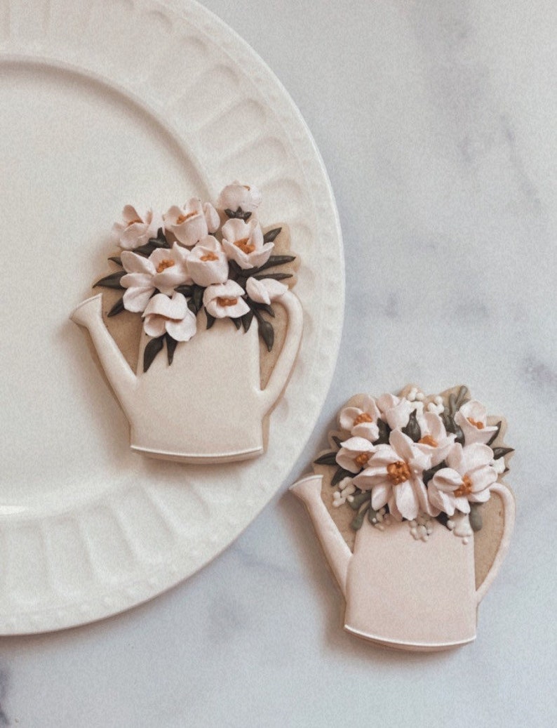 May include: Two white sugar cookies decorated with white icing and a floral design. The cookies are shaped like watering cans and are filled with white flowers. The cookies are on a white plate and a white marble surface.